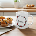 White mug with 'friend' text and floral design on a kitchen counter with muffins