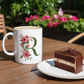 White mug with floral design and letter 'R' next to a slice of chocolate cake on a wooden table outdoors.