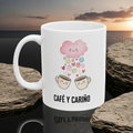 White mug with coffee-themed design and 'Café Y Cariño' text, set against a sunset over water.