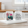 Two mugs on a wooden surface with one featuring a peacock design and the other with a romantic quote.