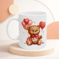 White mug with a teddy bear design holding heart balloons on a light background